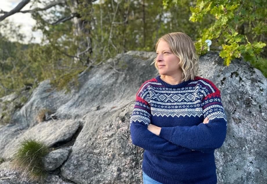 Hanne Nøding står med armene i kors foran et svaberg med furu og eik, i blå mariusgenser, og ser ut mot lyset.