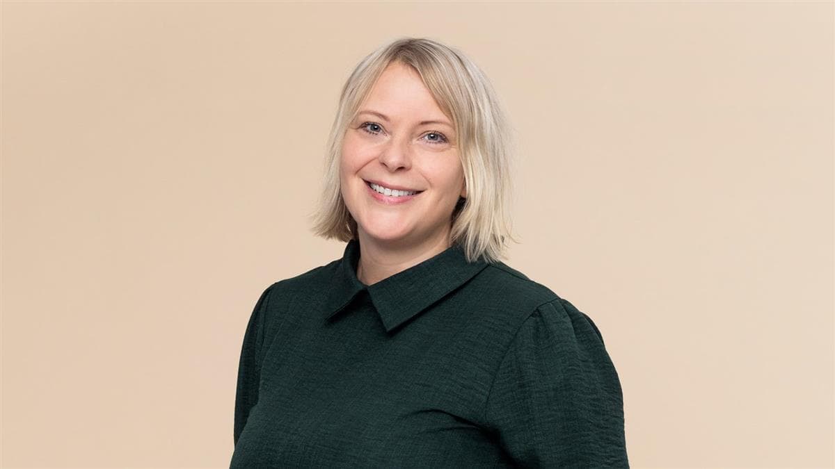 Hanne M. Nøding, portrett. Kort lyst hår, mørkegrønn bluse, beige bakgrunn.